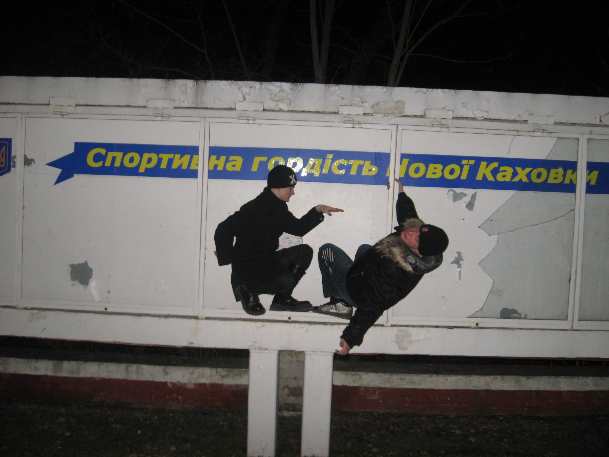 Two friends posing on a rail in front of a sporting-pride sign in Nova Kakhovka